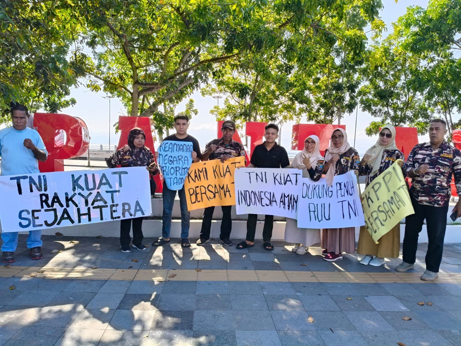 Sekelompok orang berdiri dengan spanduk yang mendukung TNI di depan tanda besar.